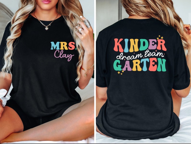 May include: Two black t-shirts with colorful text. The front shirt says "MRS. Clay" in rainbow letters. The back shirt says "Kinder dream team Garten" in rainbow letters.