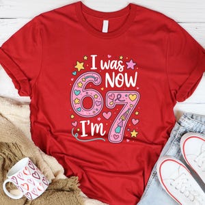 May include: Red t-shirt featuring the text "I was 6, now I'm 7" in a colourful design. The number 6 is pink with hearts and stars, and the number 7 has a unicorn horn. A fun birthday shirt.