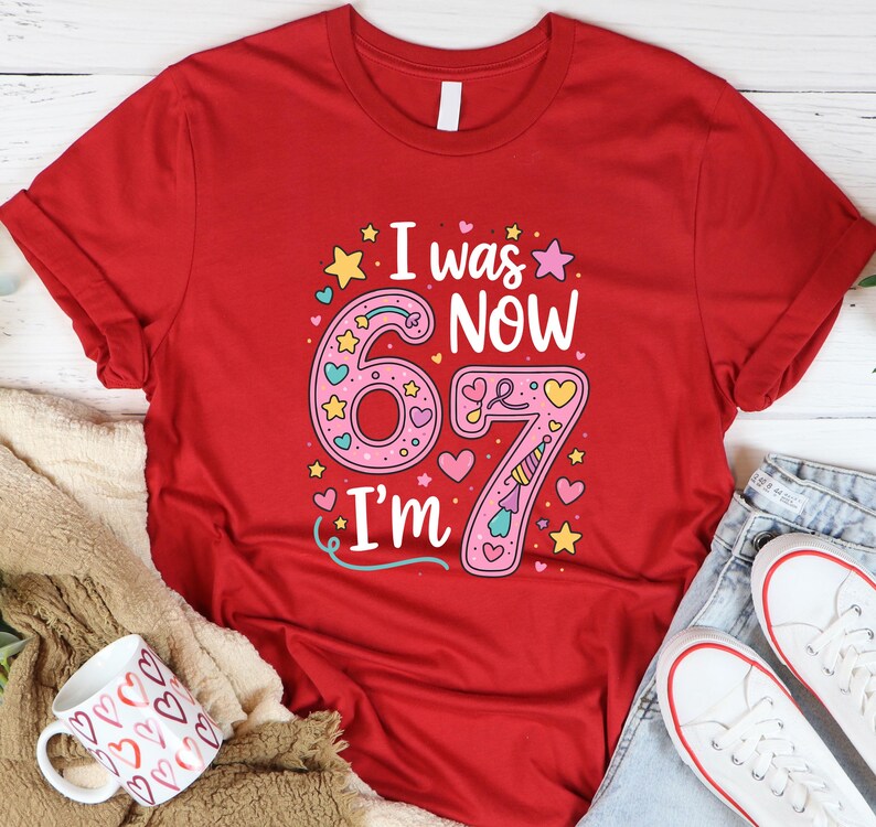 May include: Red t-shirt featuring the text "I was 6, now I'm 7" in a colourful design. The number 6 is pink with hearts and stars, and the number 7 has a unicorn horn. A fun birthday shirt.