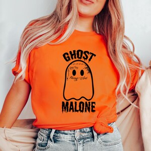 May include: Orange t-shirt featuring a black ghost design with the text "GHOST MALONE". The ghost has two oval eyes and the words "Always Tired" below it. The shirt is made of a soft material.