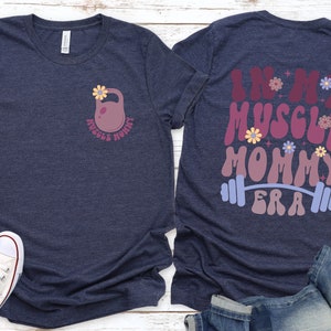 Op de afbeelding: Een marineblauw t-shirt met een roze kettlebell en bloemen aan de voorkant en de tekst "In My Muscle Mommy Era" met een halterstang en bloemen aan de achterkant.