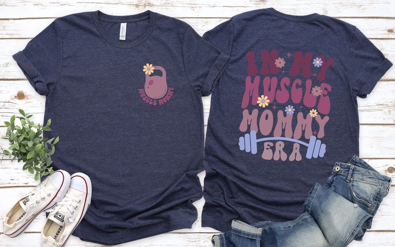 Op de afbeelding: Een marineblauw t-shirt met een roze kettlebell en bloemen aan de voorkant en de tekst "In My Muscle Mommy Era" met een halterstang en bloemen aan de achterkant.