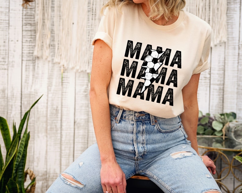 May include: A cream colored t-shirt with a black graphic of a lightning bolt made of soccer balls and the word "MAMA" repeated three times.