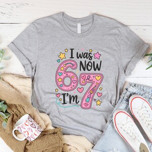 May include: A grey t-shirt with the text "I was 6 now I'm 67". The number 6 is pink with yellow stars and hearts. The number 7 is pink with a unicorn horn. The shirt is paired with jeans and white trainers.