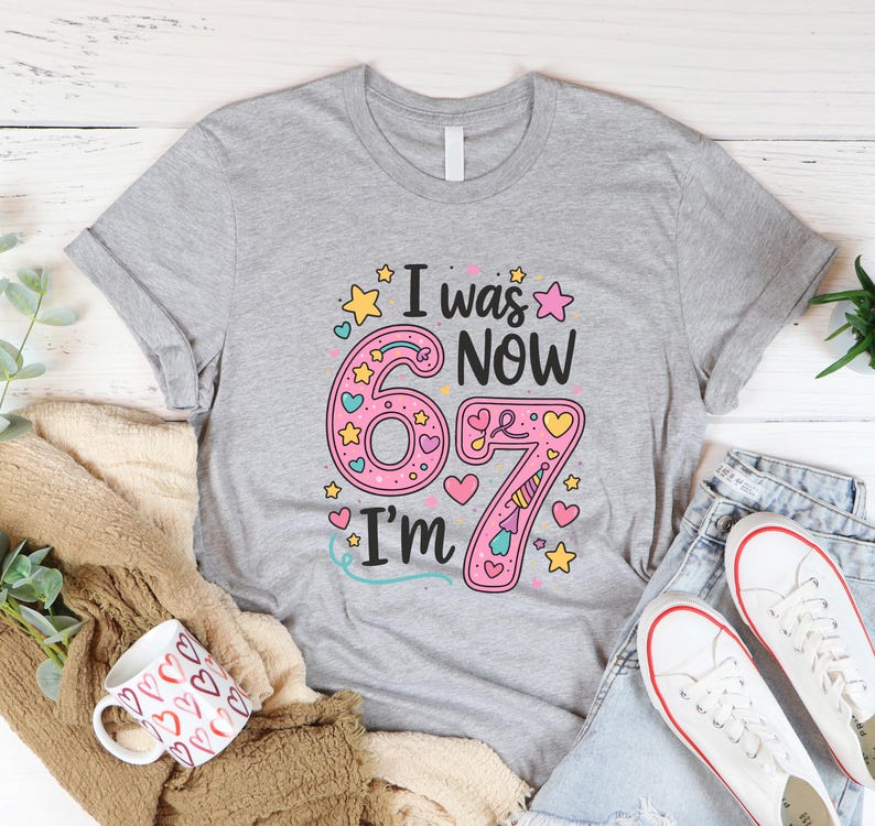 May include: A grey t-shirt with the text "I was 6 now I'm 67". The number 6 is pink with yellow stars and hearts. The number 7 is pink with a unicorn horn. The shirt is paired with jeans and white trainers.