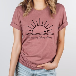 K&ouml;nnte beinhalten: Ein mauvefarbenes T-Shirt mit einer schwarzen Grafik einer Sonne, die &uuml;ber einer Wellenlinie mit einem kleinen Herzen in der Mitte aufgeht. Der Text "we rise by lifting others" ist unter der Grafik gedruckt.