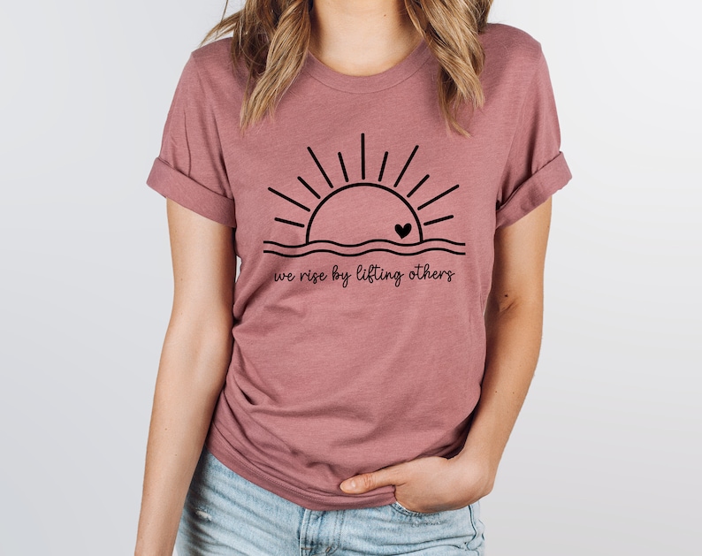 K&ouml;nnte beinhalten: Ein mauvefarbenes T-Shirt mit einer schwarzen Grafik einer Sonne, die &uuml;ber einer Wellenlinie mit einem kleinen Herzen in der Mitte aufgeht. Der Text "we rise by lifting others" ist unter der Grafik gedruckt.