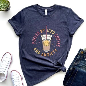 May include: A navy blue t-shirt with a graphic of a cup of iced coffee with a smiley face and the text "Fueled by Iced Coffee and Anxiety".