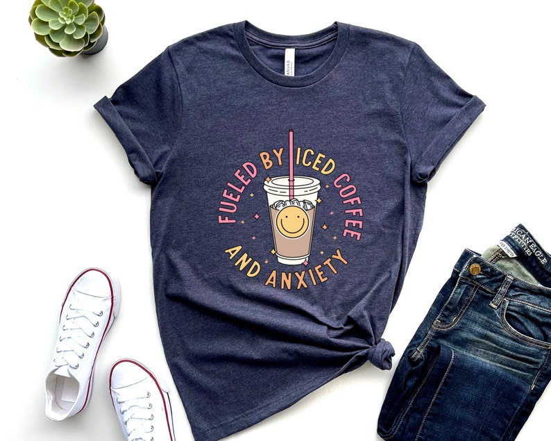 May include: A navy blue t-shirt with a graphic of a cup of iced coffee with a smiley face and the text "Fueled by Iced Coffee and Anxiety".