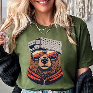 May include: Olive green t-shirt featuring a bear wearing sunglasses and a retro-style hat. The bear's sunglasses reflect a sunset scene with palm trees and a city skyline. The bear is wearing an orange jacket.