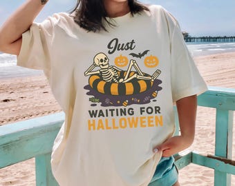 May include: Cream-colored t-shirt with a Halloween-themed graphic. The design features a skeleton relaxing on a striped floatie, pumpkins, and a bat, with the text "Just Waiting for Halloween."