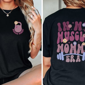 Op de afbeelding: Zwart T-shirt met een roze kettlebell en bloemen aan de voorkant en de tekst "In My Muscle Mommy Era" met een halterstang en bloemen aan de achterkant.