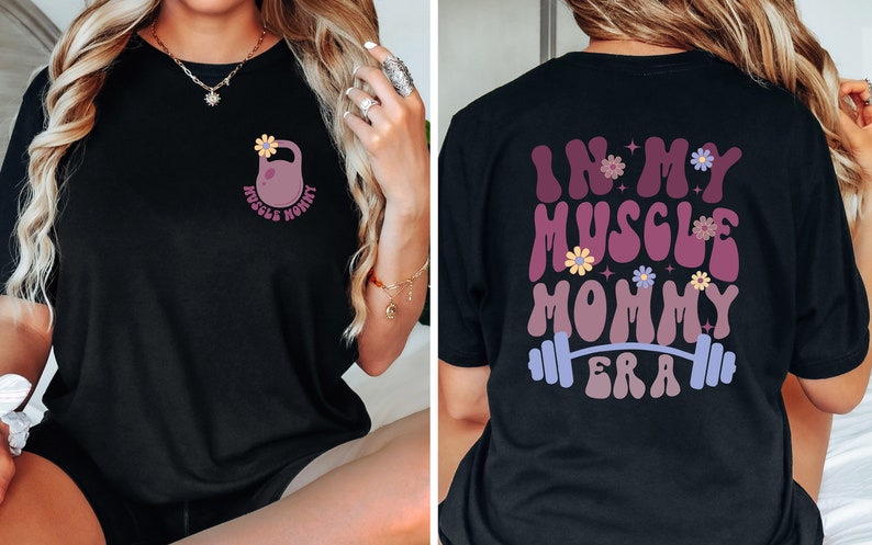 Op de afbeelding: Zwart T-shirt met een roze kettlebell en bloemen aan de voorkant en de tekst "In My Muscle Mommy Era" met een halterstang en bloemen aan de achterkant.