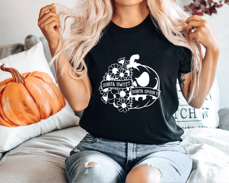 K&ouml;nnte beinhalten: Schwarzes T-Shirt mit einem wei&szlig;en Grafikdesign einer K&uuml;rbis- und Blumenkomposition. Der K&uuml;rbis-Entwurf hat den Text "Sorta Sweet Sorta Spooky" auf einem Banner. Das T-Shirt hat einen Rundhalsausschnitt.
