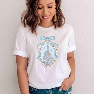 Puede incluir: Camiseta blanca con una ilustraci&oacute;n azul y blanca de un castillo con un lazo azul en la parte superior. El castillo est&aacute; rodeado por un borde de perlas.