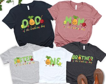 May include: A collection of colorful t-shirts with fruit-themed graphics. Designs include "DAD", "MOM", "SISTER", "ONE", and "BROTHER" with a pineapple and other fruit characters. The shirts are in various colors, including navy, pink, gray, and white.