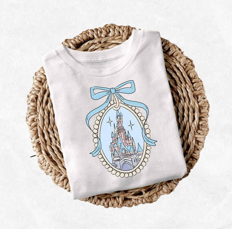Puede incluir: Camiseta blanca con un lazo azul y un marco engastado con perlas que contiene una ilustraci&oacute;n de dibujos animados de un castillo rosa y azul.