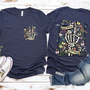May include: Two navy blue t-shirts featuring a skeleton hand making a peace sign surrounded by flowers and butterflies. The text on the shirt reads "Good energy is contagious".