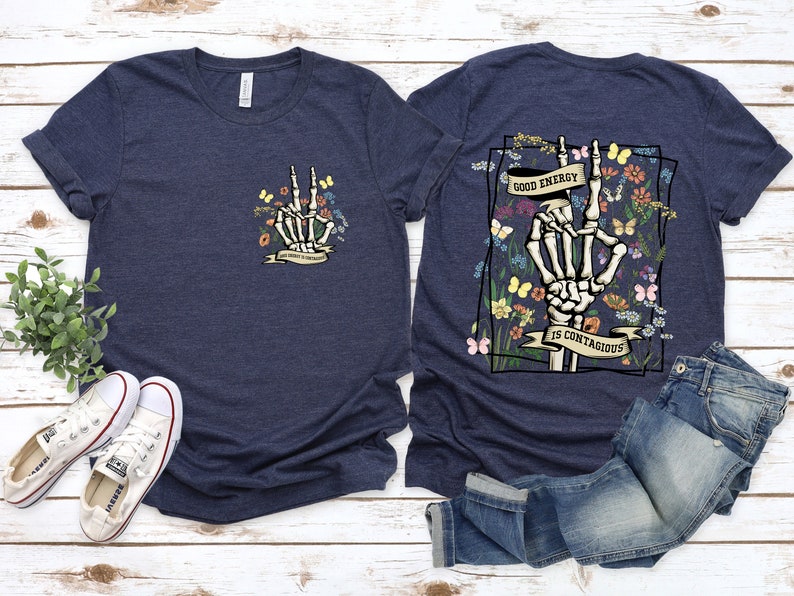 May include: Two navy blue t-shirts featuring a skeleton hand making a peace sign surrounded by flowers and butterflies. The text on the shirt reads "Good energy is contagious".