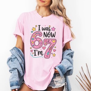 May include: A pale pink t-shirt featuring the phrase "I was 6, now I'm 7". The number 6 is large and pink, and the number 7 is pink with a unicorn horn. The shirt has a round neck and short sleeves.