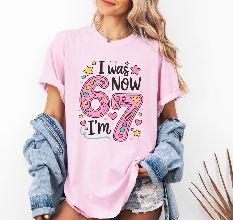 May include: A pale pink t-shirt featuring the phrase "I was 6, now I'm 7". The number 6 is large and pink, and the number 7 is pink with a unicorn horn. The shirt has a round neck and short sleeves.