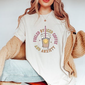 May include: White t-shirt with a graphic of a cup of iced coffee with a smiley face and the text "Fueled by Iced Coffee and Anxiety".