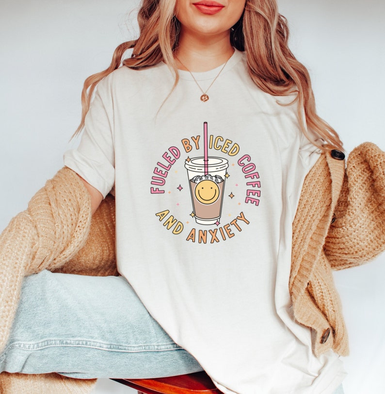 May include: White t-shirt with a graphic of a cup of iced coffee with a smiley face and the text "Fueled by Iced Coffee and Anxiety".