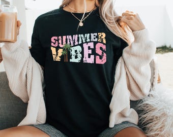 May include: Black crew neck t-shirt with the text "SUMMER VIBES" in a colorful, distressed font. The design includes a palm tree graphic. The shirt is worn with a cream-colored cardigan and denim shorts.