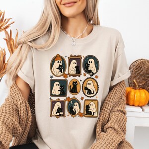 Puede incluir: Camiseta beige con ilustraciones enmarcadas de fantasmas y gatos negros. La obra de arte incluye varios estilos de marcos, hojas de oto&ntilde;o y una calabaza. La camiseta tiene un corte holgado y mangas cortas, ideal para Halloween o el oto&ntilde;o.
