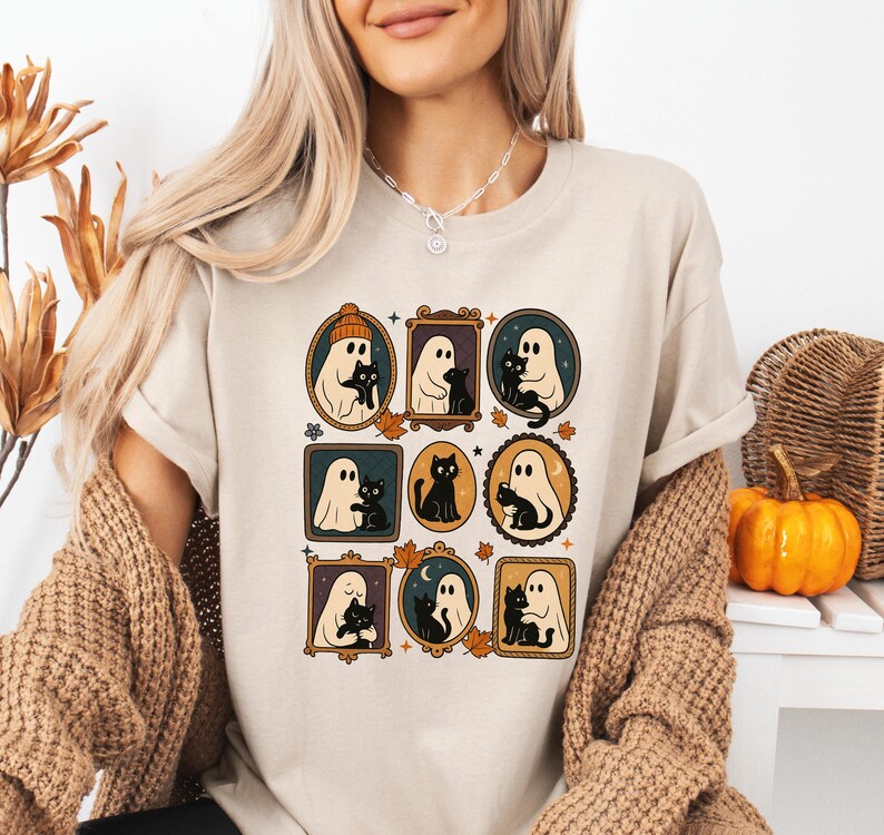 Puede incluir: Camiseta beige con ilustraciones enmarcadas de fantasmas y gatos negros. La obra de arte incluye varios estilos de marcos, hojas de oto&ntilde;o y una calabaza. La camiseta tiene un corte holgado y mangas cortas, ideal para Halloween o el oto&ntilde;o.