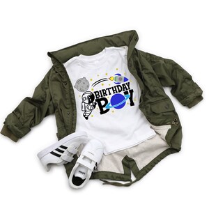 May include: White t-shirt featuring a black and blue astronaut graphic and the text "Birthday Boy". The shirt is on top of a green jacket and a pair of white trainers.