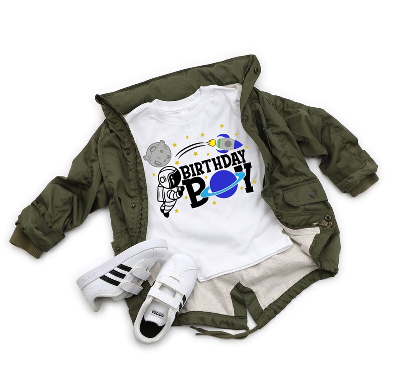 May include: White t-shirt featuring a black and blue astronaut graphic and the text "Birthday Boy". The shirt is on top of a green jacket and a pair of white trainers.
