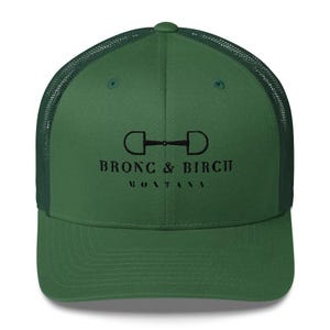 May include: A green trucker hat with a mesh back and a curved brim. The front of the hat features the text "BRONC & BIRCH MONTANA" in black, along with a graphic of a horse bit.