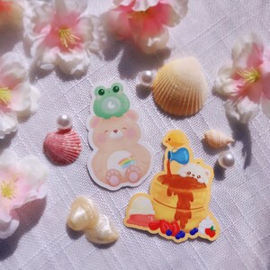 May include: Two illustrated stickers featuring a cartoon bear wearing a frog hat and a pancake stack with a cartoon chick pouring syrup. The stickers are on a white surface with pink flowers and seashells.
