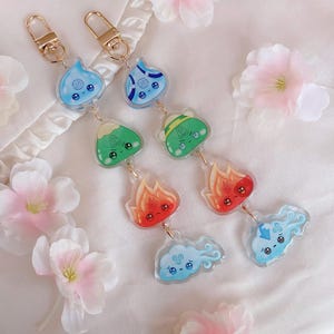 Puede incluir: Dos llaveros de oro con dijes que representan los cuatro elementos. Los dijes son agua, tierra, fuego y aire. El dije de agua es una lágrima azul con una cara, el dije de tierra es un triángulo verde con una cara, el dije de fuego es una llama roja con una cara, y el dije de aire es una nube azul con una cara.