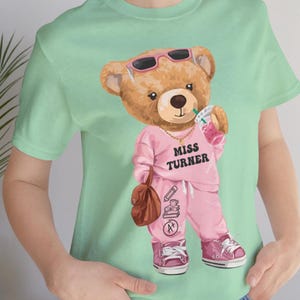 May include: A light green t-shirt featuring a cartoon teddy bear wearing sunglasses and a pink sweatsuit with the text "MISS TURNER". The bear is holding a drink and a brown satchel. The bear's outfit includes a gold necklace and pink sneakers.