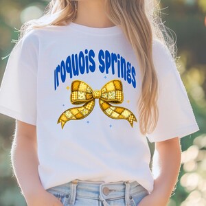 May include: White t-shirt featuring the text "Iroquois Springs" in blue, arched above a yellow bow graphic. The bow has a patchwork design with yellow and white squares. Small blue star accents are also present.