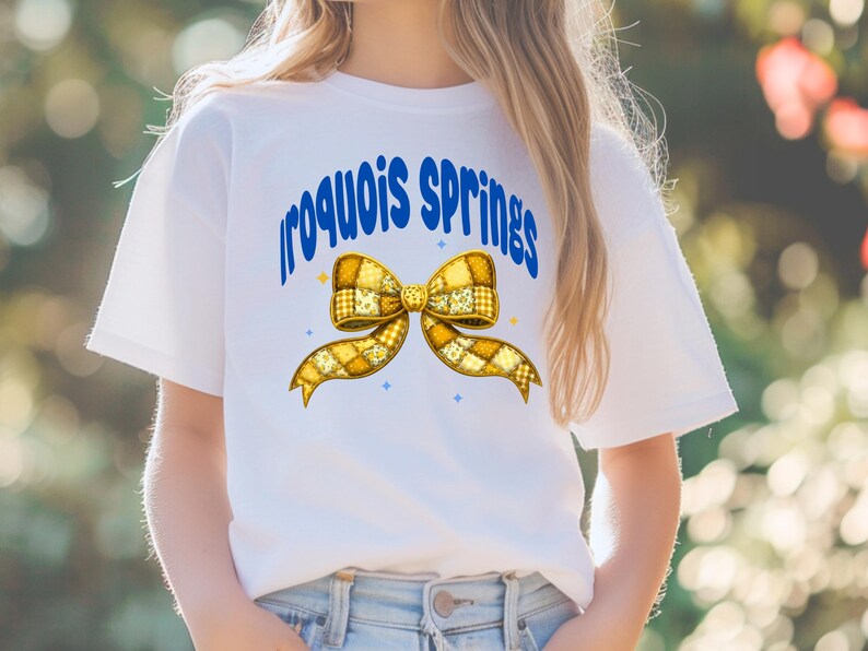 May include: White t-shirt featuring the text "Iroquois Springs" in blue, arched above a yellow bow graphic. The bow has a patchwork design with yellow and white squares. Small blue star accents are also present.