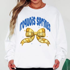 May include: White sweatshirt with the text "Iroquois Springs" in blue arched lettering. A large yellow bow with a patchwork design is centered below the text. The bow has gold and yellow tones with blue star accents.