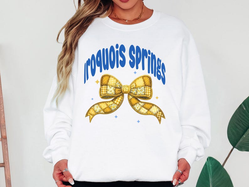 May include: White sweatshirt with the text "Iroquois Springs" in blue, arched above a yellow patchwork bow. The bow has a detailed design with various shades of yellow and gold. Small blue stars are scattered around the bow.