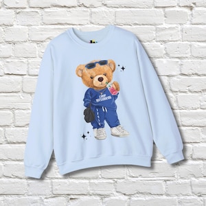May include: Light blue sweatshirt featuring a cartoon bear wearing sunglasses and a blue tracksuit, holding a drink. The bear has the text "Camp Westmoon" on its outfit. The sweatshirt is on a white brick wall.