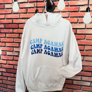 May include: White hooded sweatshirt with a blue and white graphic that says "Camp Agawak" in a retro font.