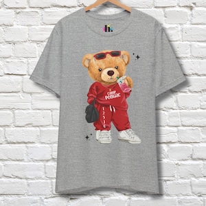 May include: A gray t-shirt featuring a cartoon teddy bear wearing sunglasses and a red tracksuit, holding a drink. The bear has a black bag and white sneakers. The text "Camp Pontiac" is on the shirt.