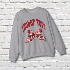 May include: A light gray sweatshirt with the text "Timber Tops" in red, arched across the top. A patchwork bow design in red, pink, and white is centered below the text. The sweatshirt is displayed against a white brick wall.