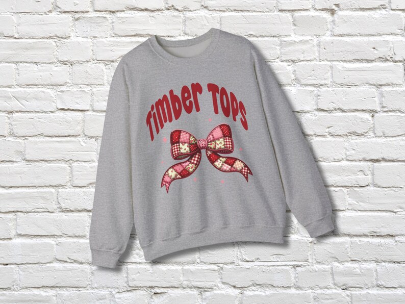 May include: A light gray sweatshirt with the text "Timber Tops" in red, arched across the top. A patchwork bow design in red, pink, and white is centered below the text. The sweatshirt is displayed against a white brick wall.