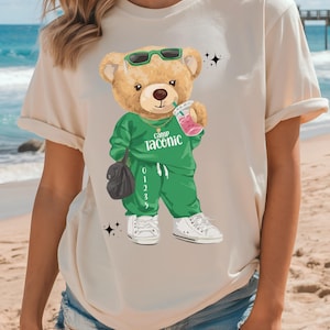 May include: Beige t-shirt featuring a cartoon teddy bear wearing sunglasses and a green tracksuit with the text "Camp Taconic." The bear is holding a pink drink and a black bag, with white sneakers. The shirt is on a person at the beach.