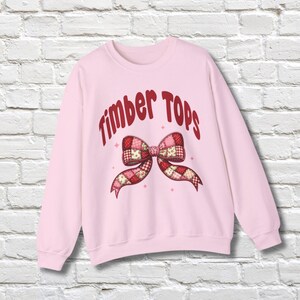 May include: A light pink sweatshirt with the text "Timber Tops" in red script above a patchwork bow design. The bow features red, pink, and white patterns. The sweatshirt is displayed against a white brick wall.