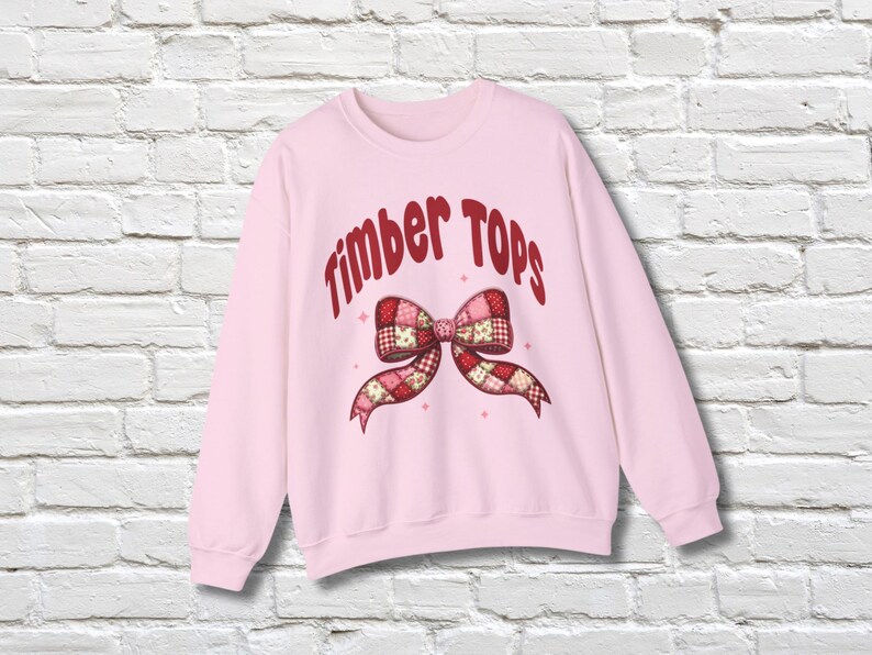 May include: A light pink sweatshirt with the text "Timber Tops" in red script above a patchwork bow design. The bow features red, pink, and white patterns. The sweatshirt is displayed against a white brick wall.