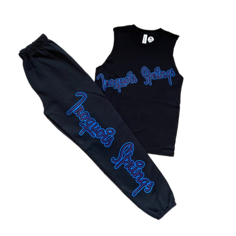 Puede incluir: Pantalones de ch&aacute;ndal negros con un gr&aacute;fico "Inoquois Springs" azul y morado en la pierna y una camiseta sin mangas negra con el mismo gr&aacute;fico en la parte delantera.