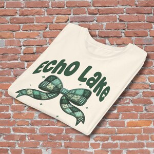 May include: Cream-colored t-shirt featuring the text "Echo Lake" in dark green, with a plaid bow graphic in shades of green and teal. The shirt is folded and displayed against a brick wall.